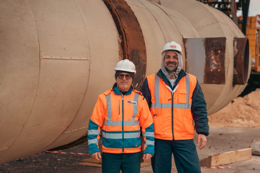 pexels-photo-36673082-36673082 Two construction workers in safety gear standing by industrial machinery on a construction site.